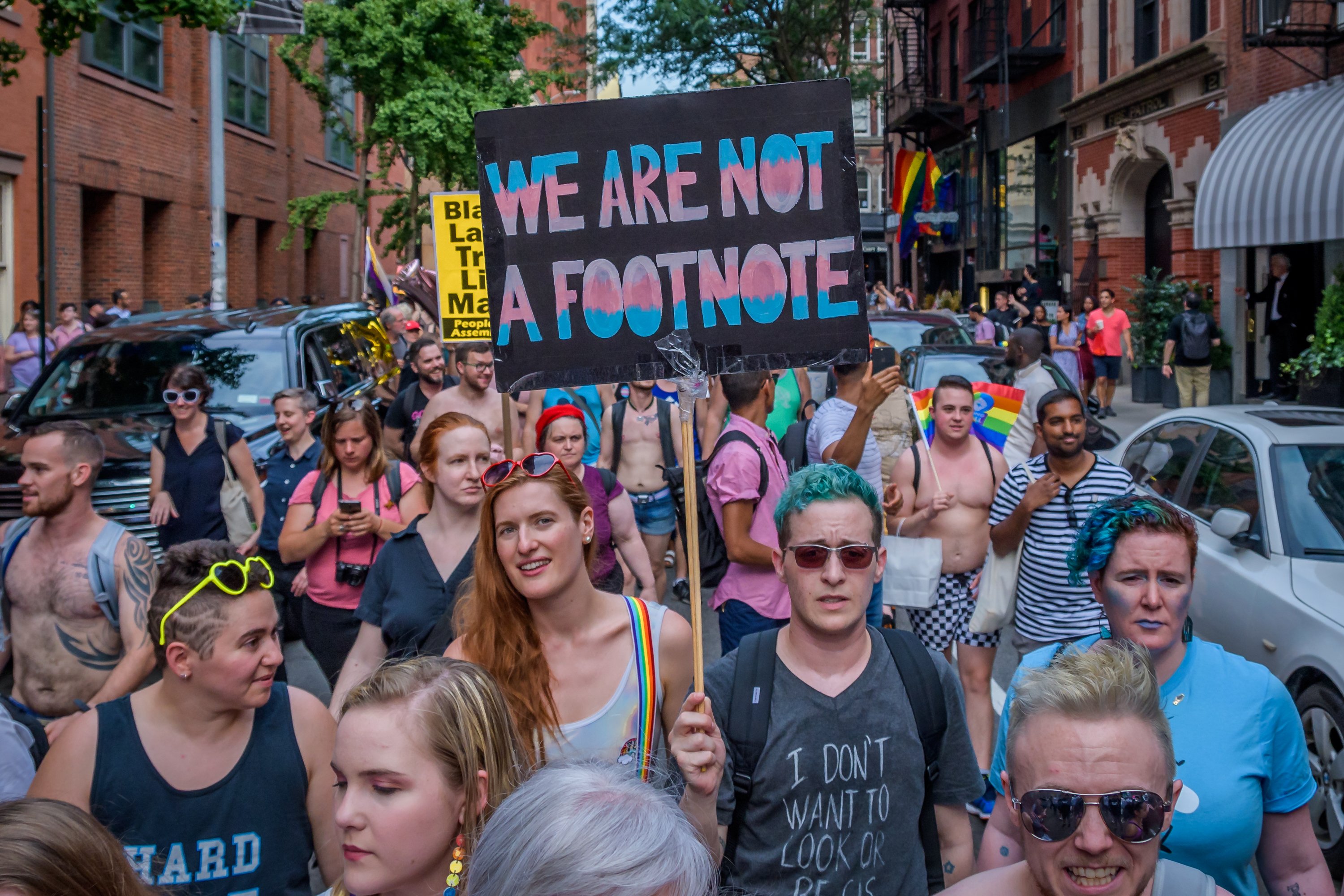 Image of a large group of diverse people marching on a city street holding LGBTQ+ allyship signs including one that says "WE ARE NOT A FOOTNOTE" in pink, blue, and white.